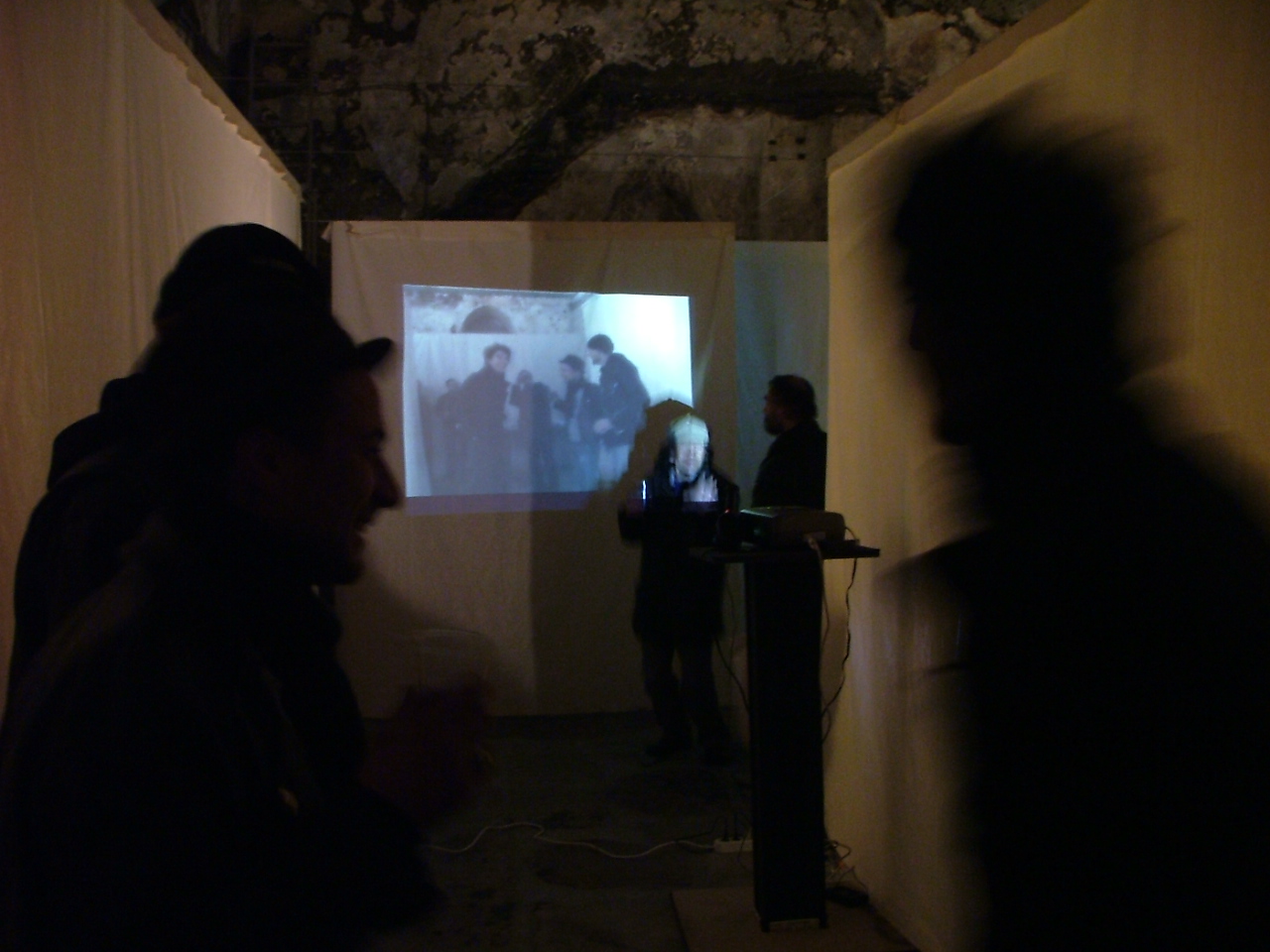 Exhibition visitors walking past a digital projector casting a video loop onto a white fabric wall inside a stone-vaulted hall.